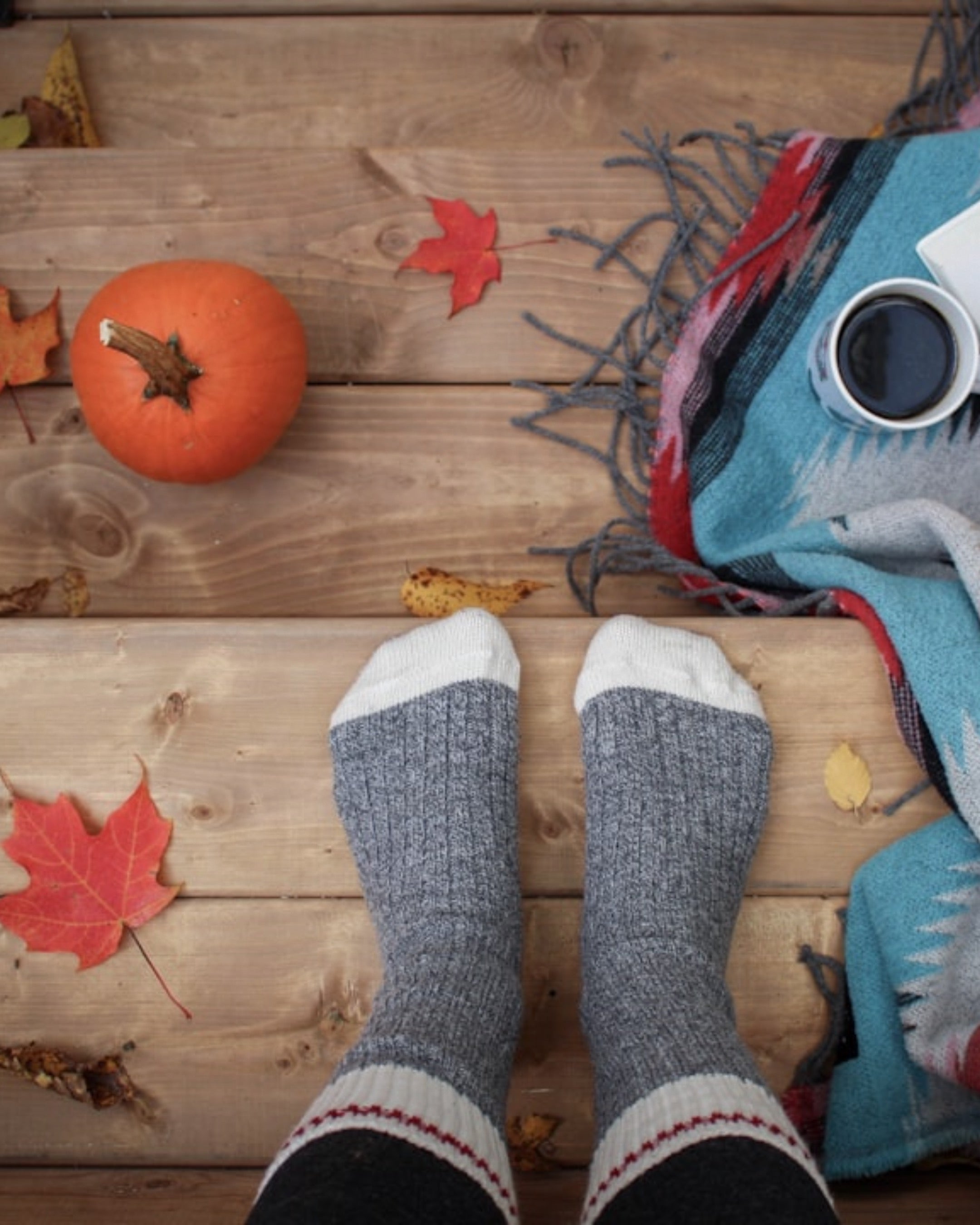 Image of someone sitting outside with a fall scene including a small pumpkin, warm blanket, and hot coffee