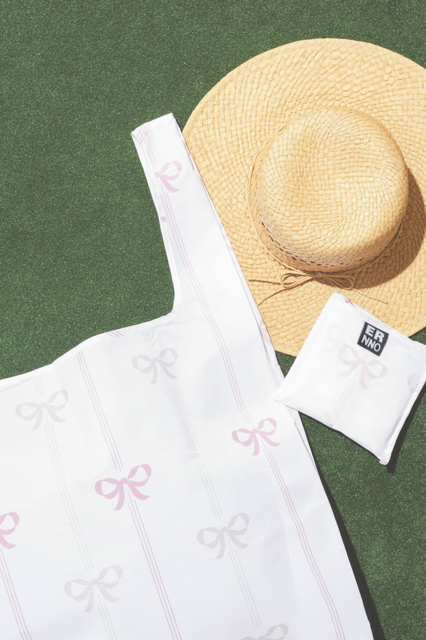 A white tote bag with pink and cream bow accents, displayed alongside a matching white object and a straw hat on a grass-like surface.