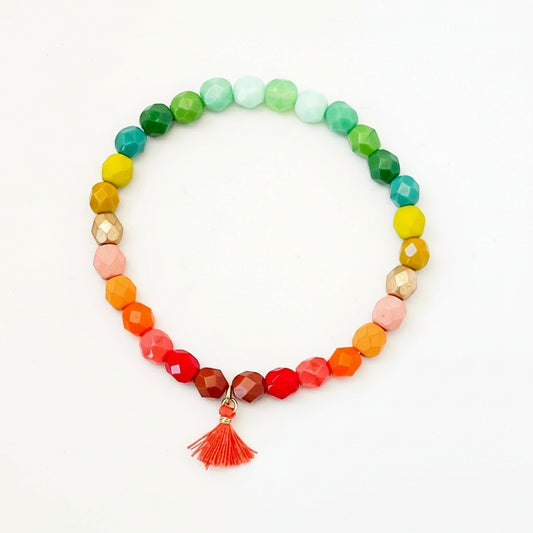 A multicolored rainbow ombre beaded bracelet with a red cotton tassel, made of Czech glass, displayed against a white background.