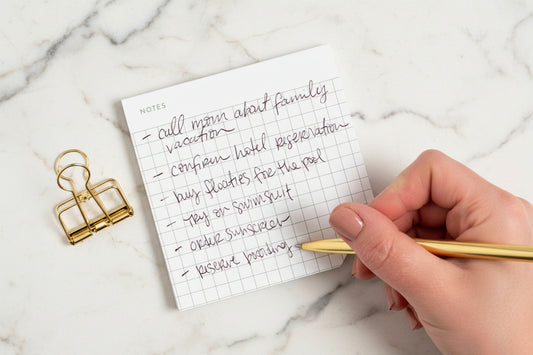 Handwritten to-do list on a small notepad with a gold pen and clip on a white background