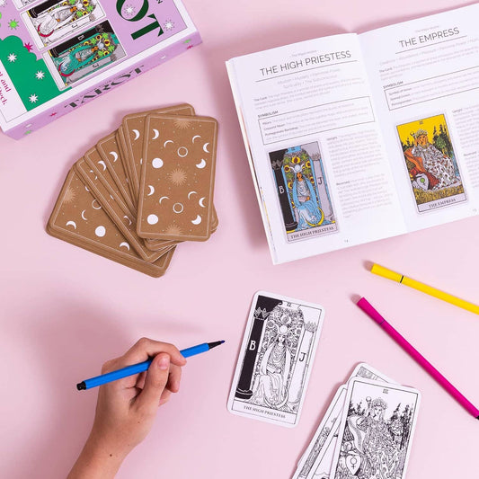 Tarot cards and book on a pink surface with a hand holding a pen.