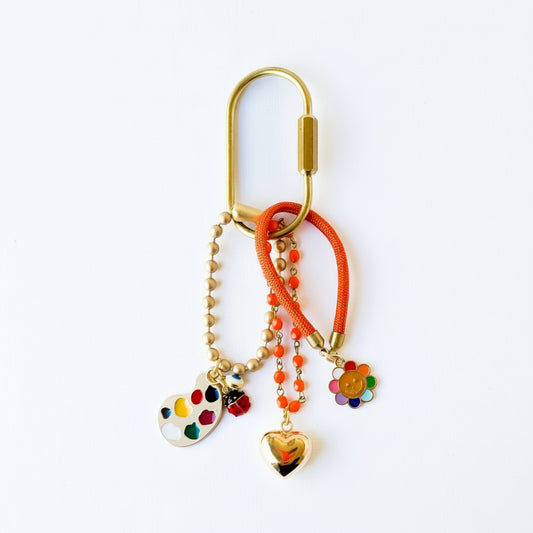 Close-up of keychain with gold heart, smiley face, rainbow, evil eye and ladybug