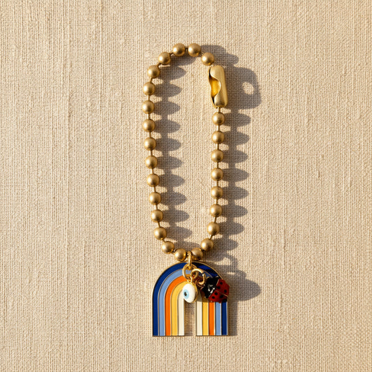 A gold ball chain bag charms with a multicolored rainbow charms and small enameled figures on a beige textured background.