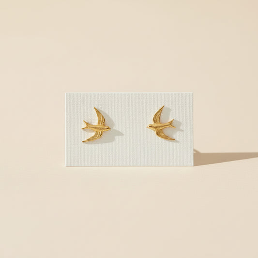 Gold bird-shaped earrings on a light surface with a blurred background of shelves and bags.
