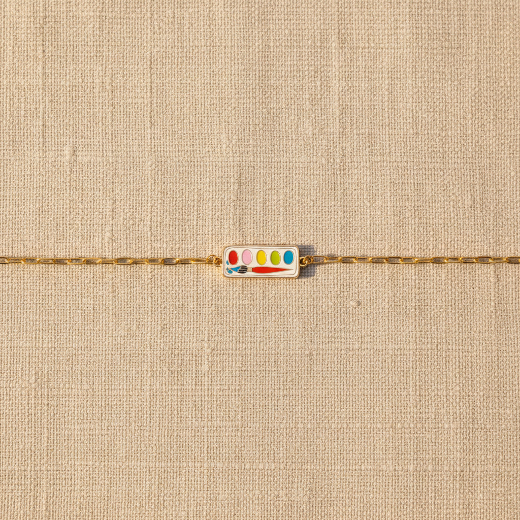 Gold bracelet with a colorful paint palette charm on a beige textured background