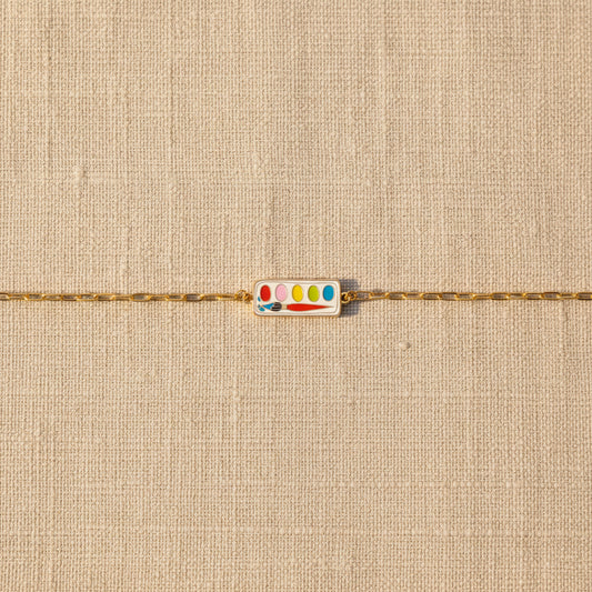 Gold bracelet with a colorful paint palette charm on a beige textured background