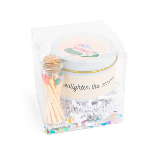 Birthday cake scented candle in white tin with corked vial of colorful matchsticks, packaged in a clear vinyl gift box.