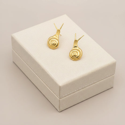 Gold snail-shaped earrings held in a hand against a textured gray background