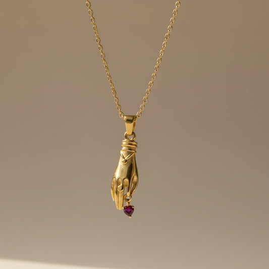 Gold necklace with a hand and heart pendant held in a hand against a gray background