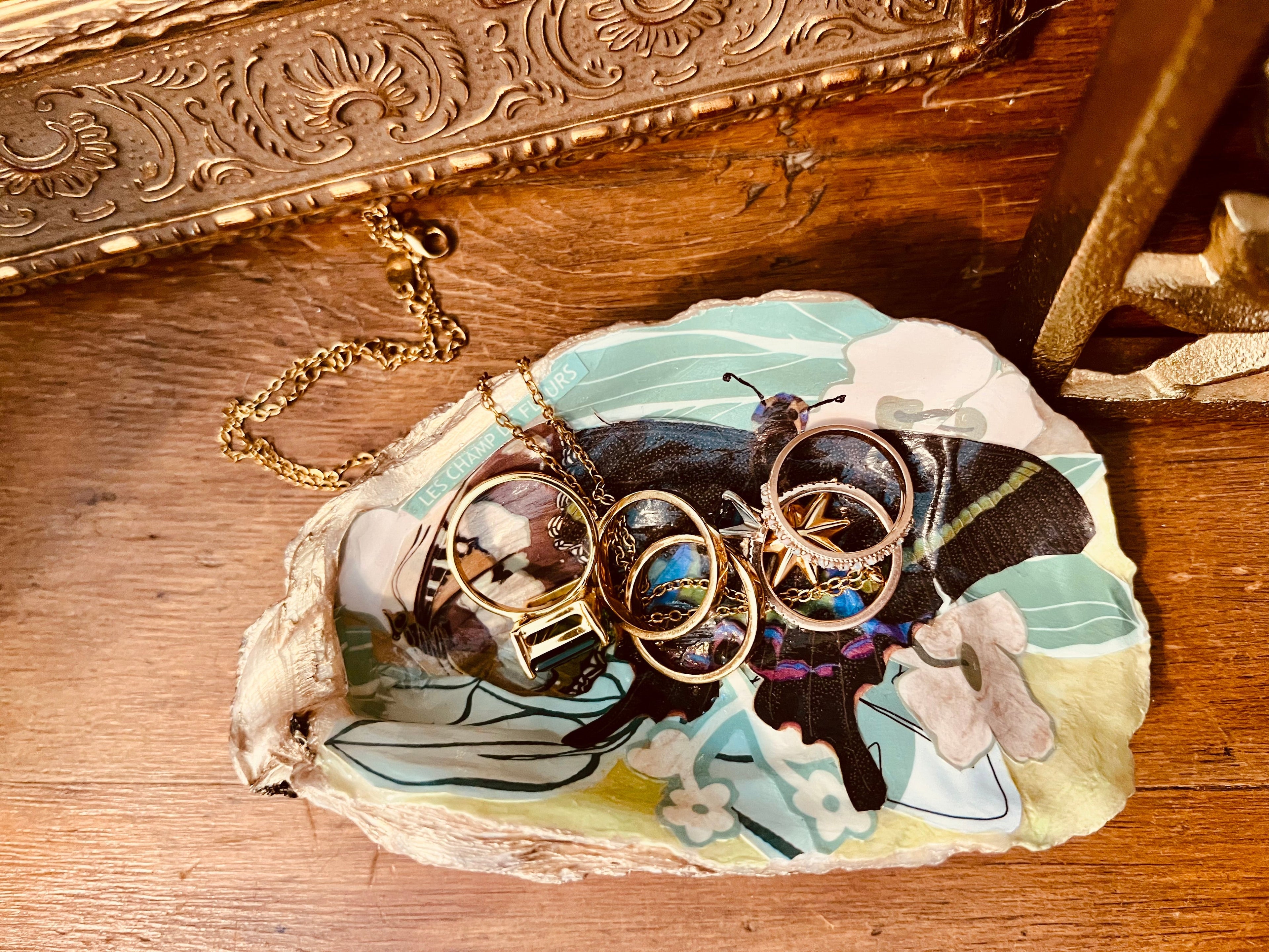 A handcrafted oyster shell trinket dish with floral and butterfly collage, gold accents, and glossy finish, perfect for holding jewelry. Created by Rochambeau Home