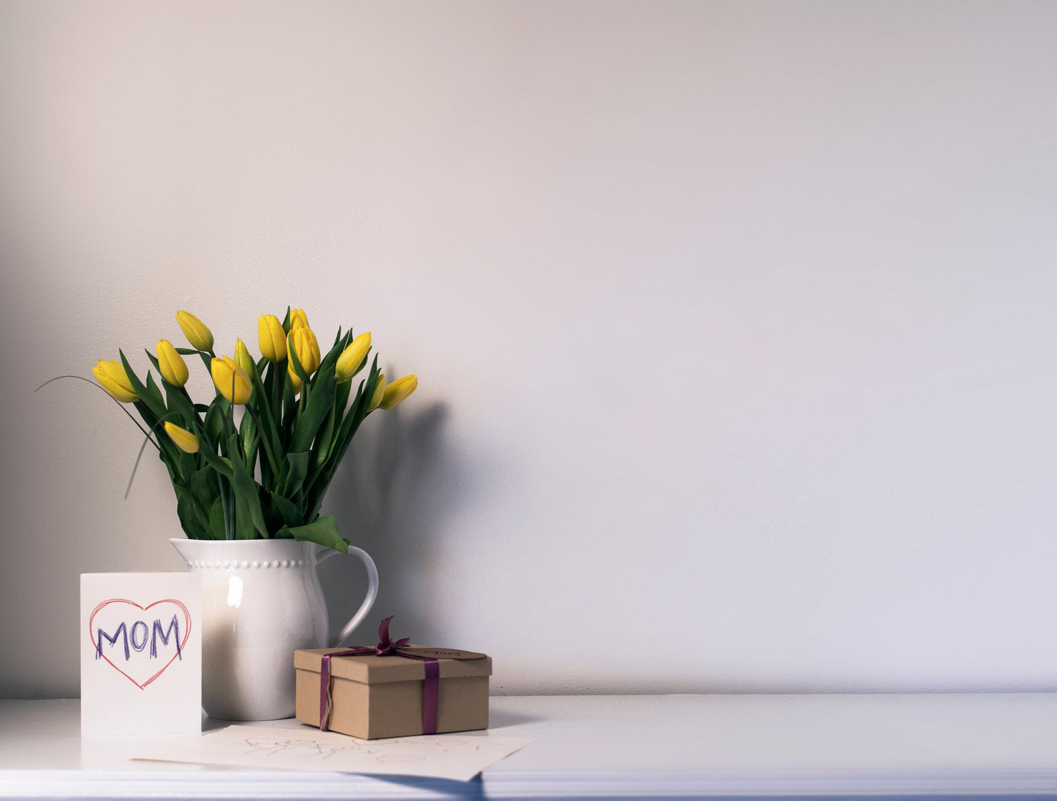 Small gift box next to a vase of yellow tulips and a card with 'MOM' surrounded by a heart.