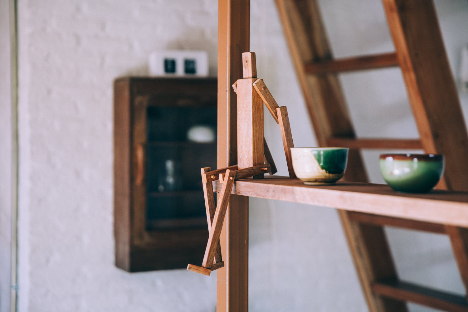 A wooden stick figure on a wooden ledge with small artisan pottery items and vintage details in the background of a white wall
