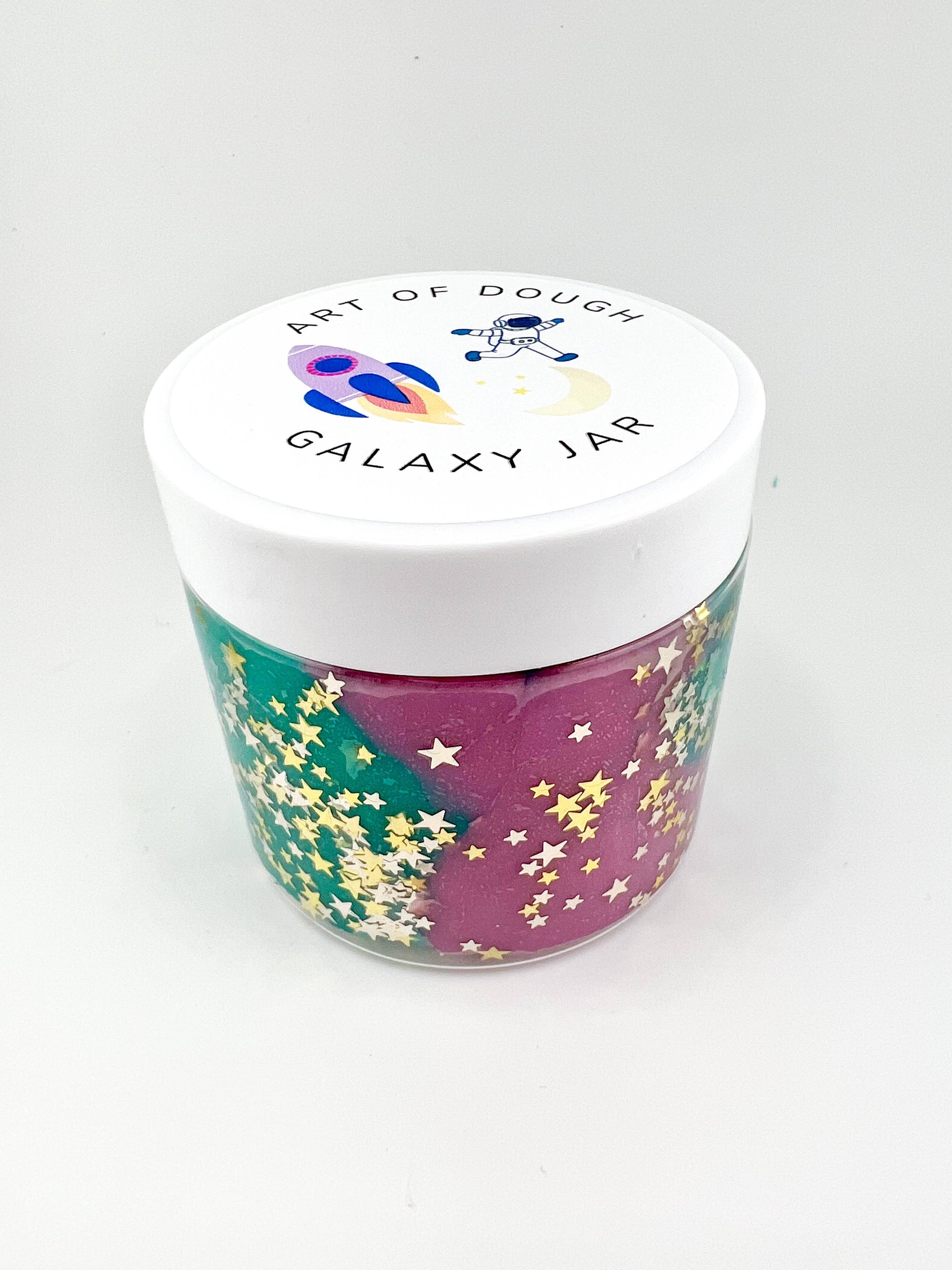 A 6oz jar with star-glitter sensory dough, a rocket ship cutter, a space figurine, and assorted gems for sensory engagement.