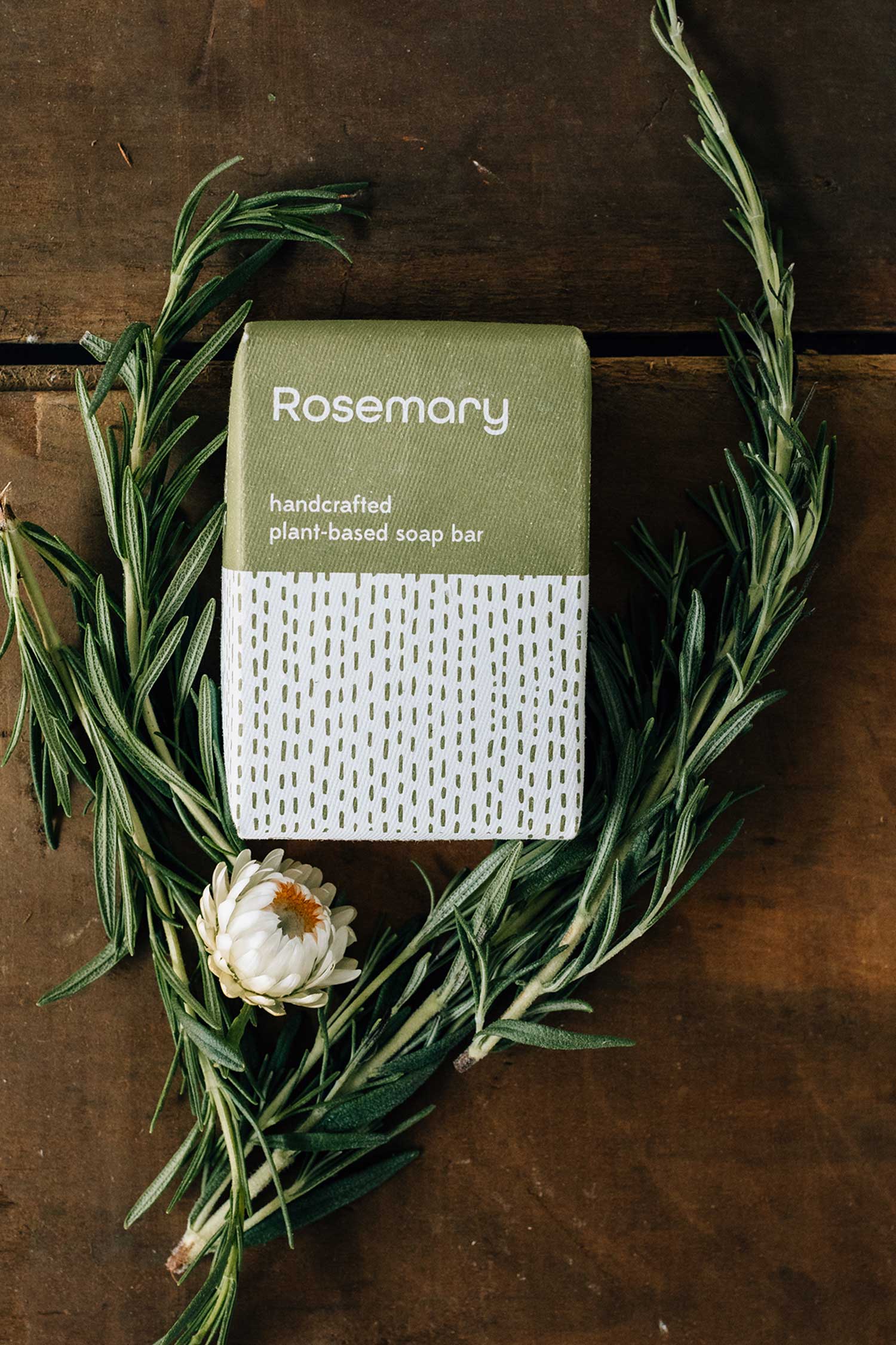 Rosemary soap bar with packaging on a wooden surface with rosemary leaves and a flower.