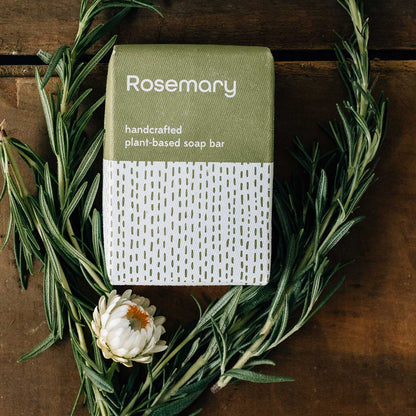 Rosemary soap bar with packaging on a wooden surface with rosemary leaves and a flower.