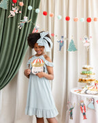 Young girl in a light blue dress with a decorative headband standing in front of a festive backdrop with decorations.
