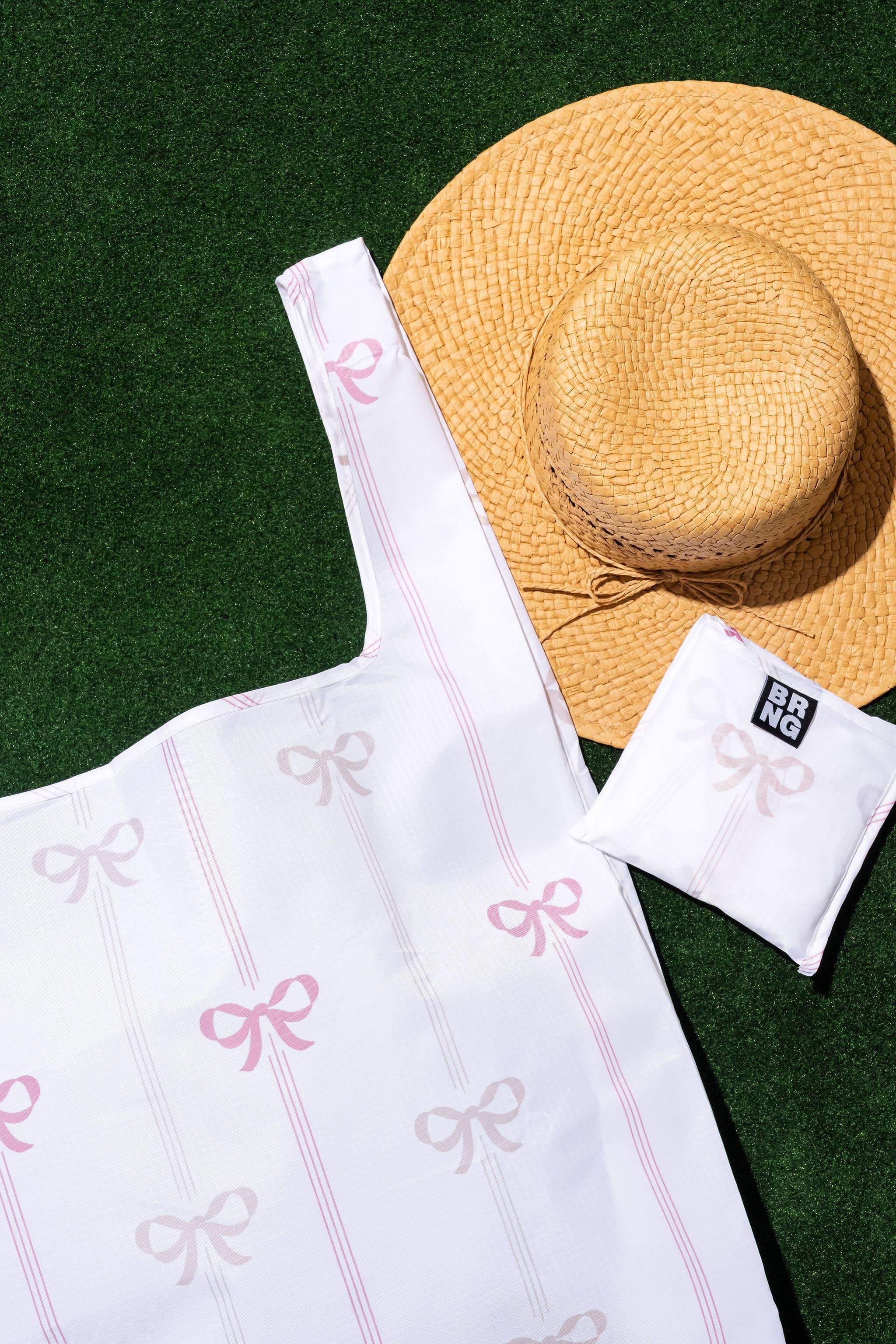 A white tote bag with pink and cream bow accents, displayed alongside a matching white object and a straw hat on a grass-like surface.