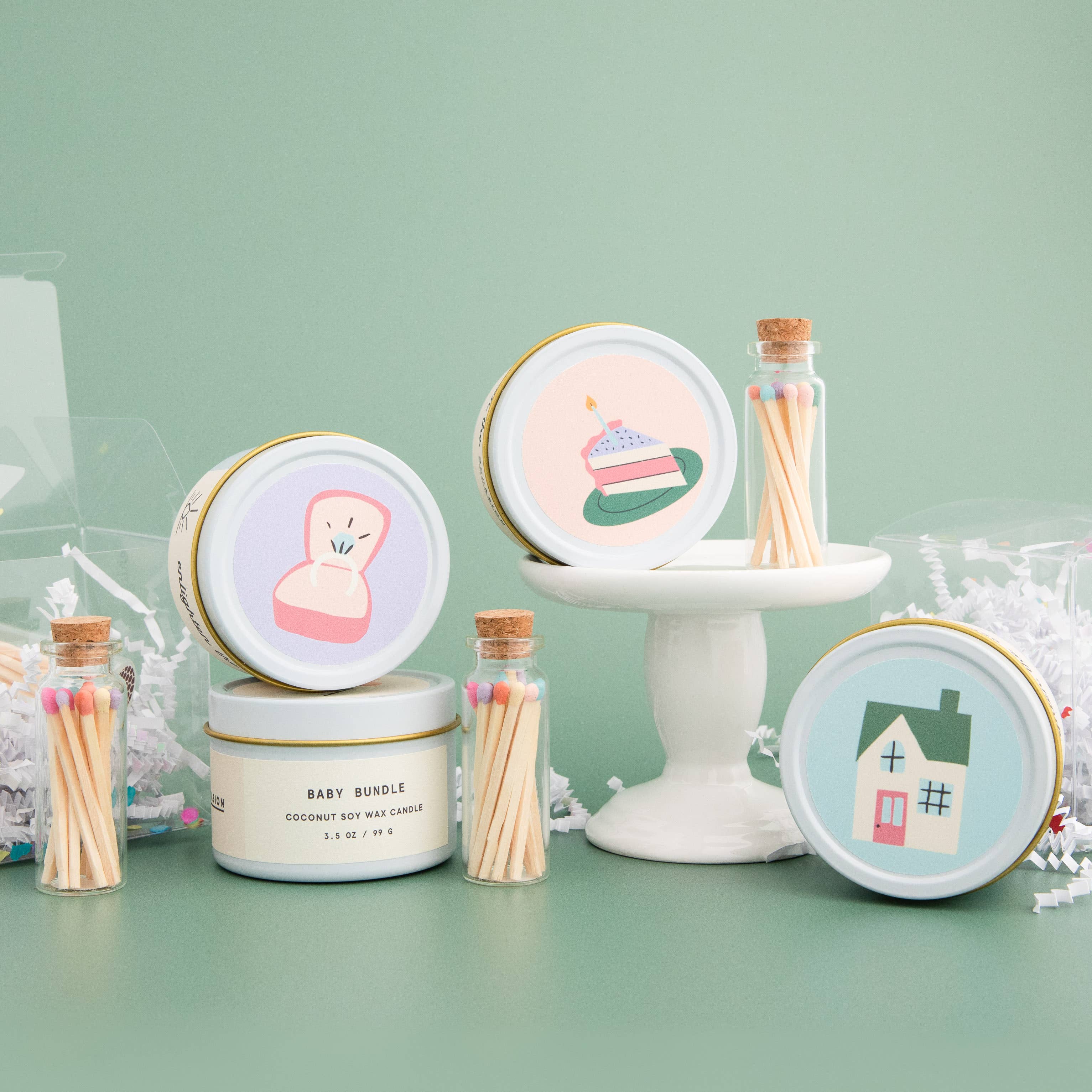 Wedding cake scented candle in white tin with corked vial of colorful matchsticks, packaged in a clear vinyl gift box.