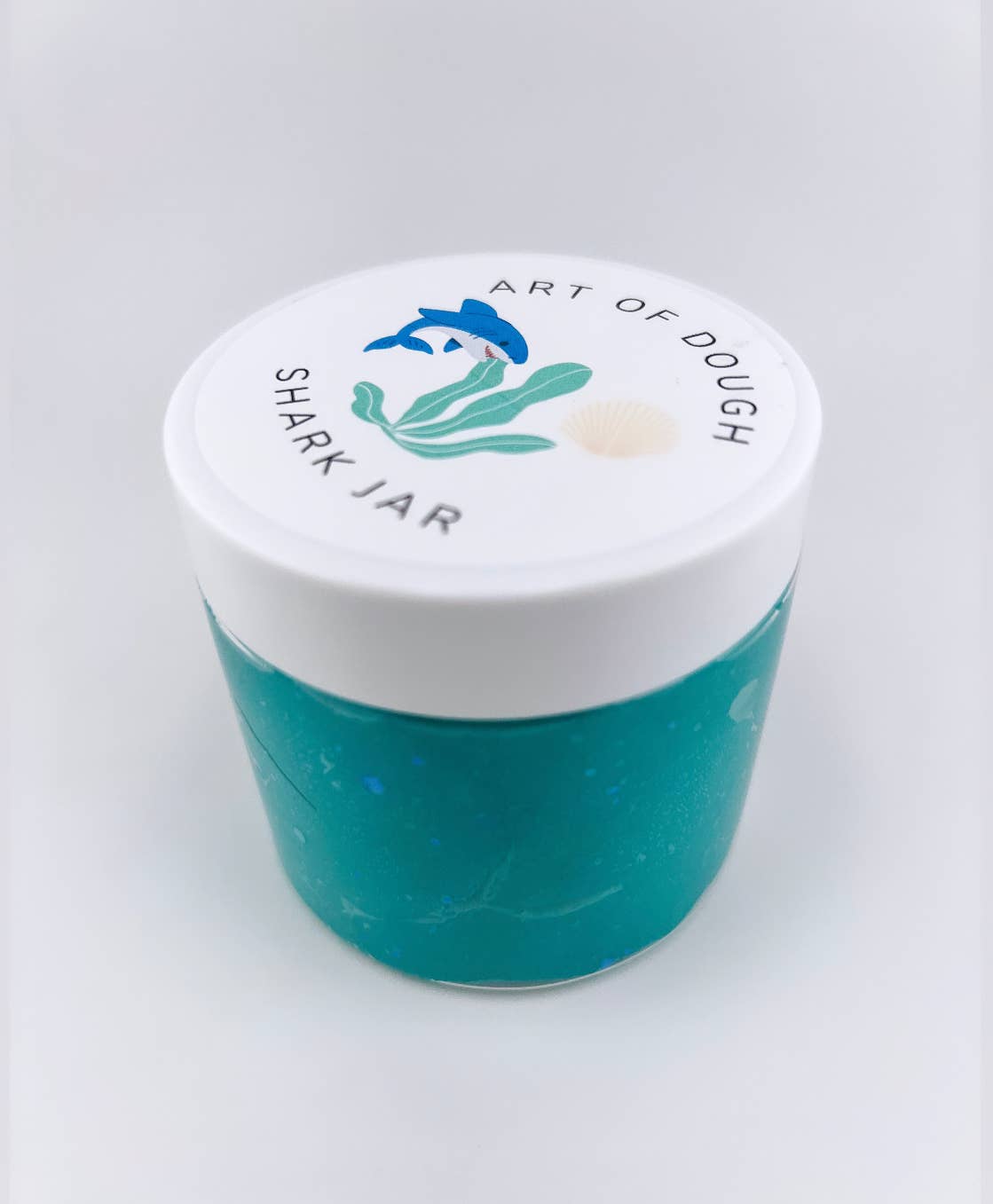 A close up of the backside of the 6oz jar containing blue playdough, a shark figurine, a faux plant, seashells, and gems, ideal for sensory play.