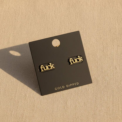 Gold 'fuck' earrings on a black card with 'Gold Dipped' text, set against a beige background.