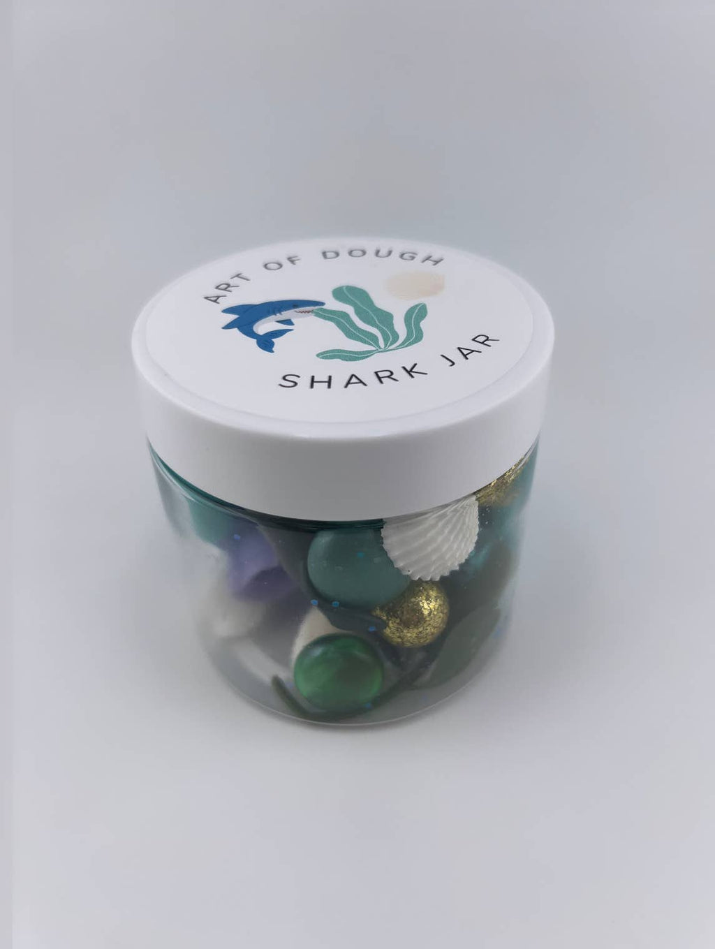 A 6oz jar containing blue playdough, a shark figurine, a faux plant, seashells, and gems, ideal for sensory play."