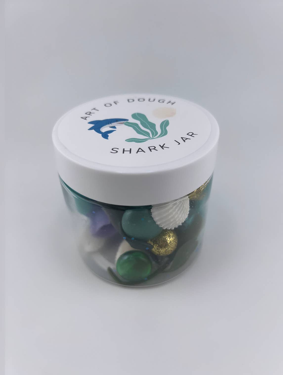 A 6oz jar containing blue playdough, a shark figurine, a faux plant, seashells, and gems, ideal for sensory play."