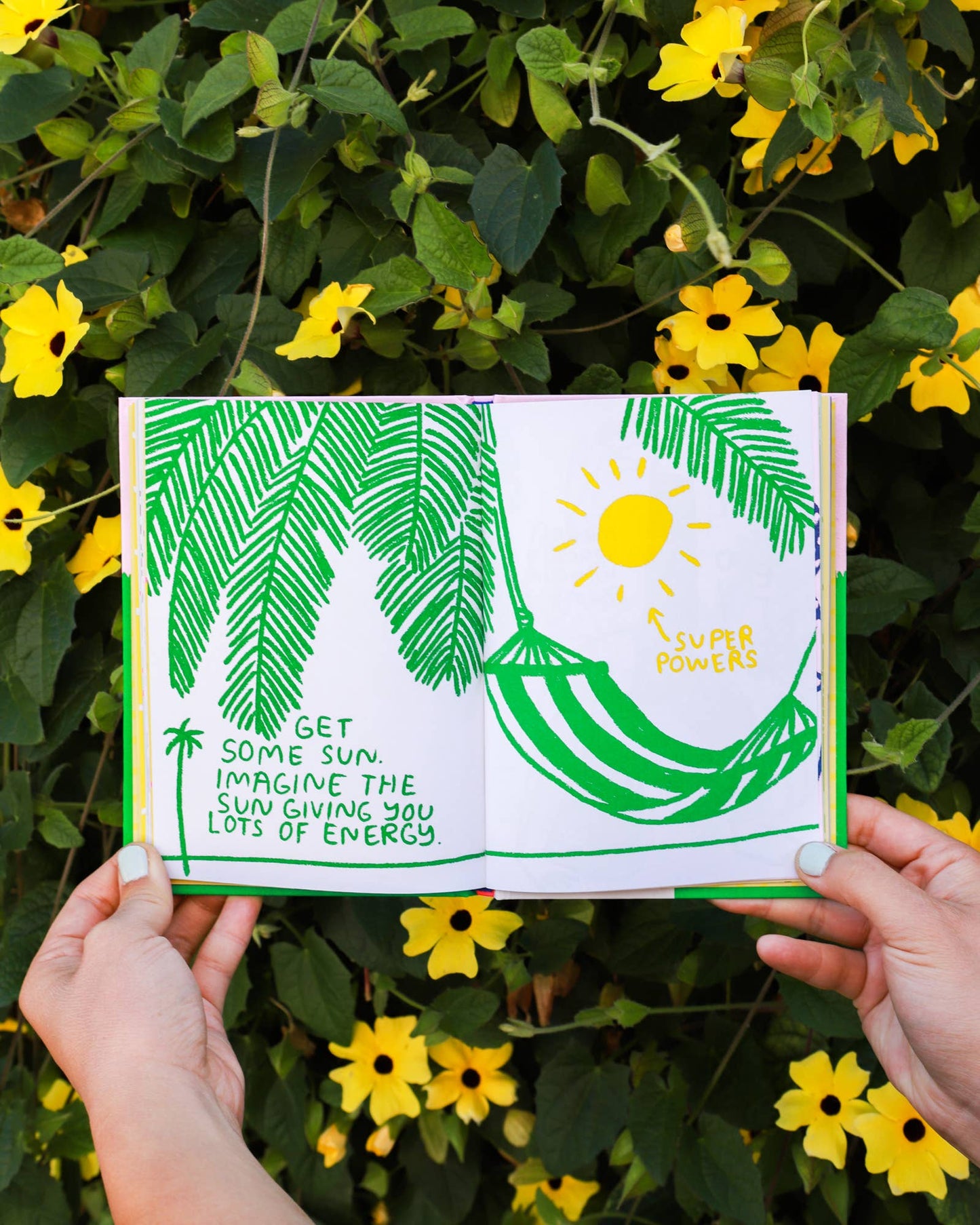 Illustrated book with green leaves and sun design held by hands against a natural background with yellow flowers.