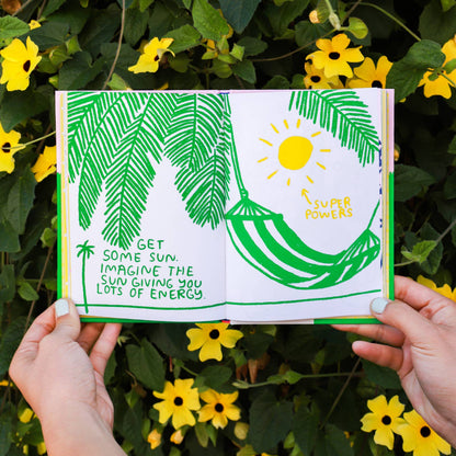 Illustrated book with green leaves and sun design held by hands against a natural background with yellow flowers.