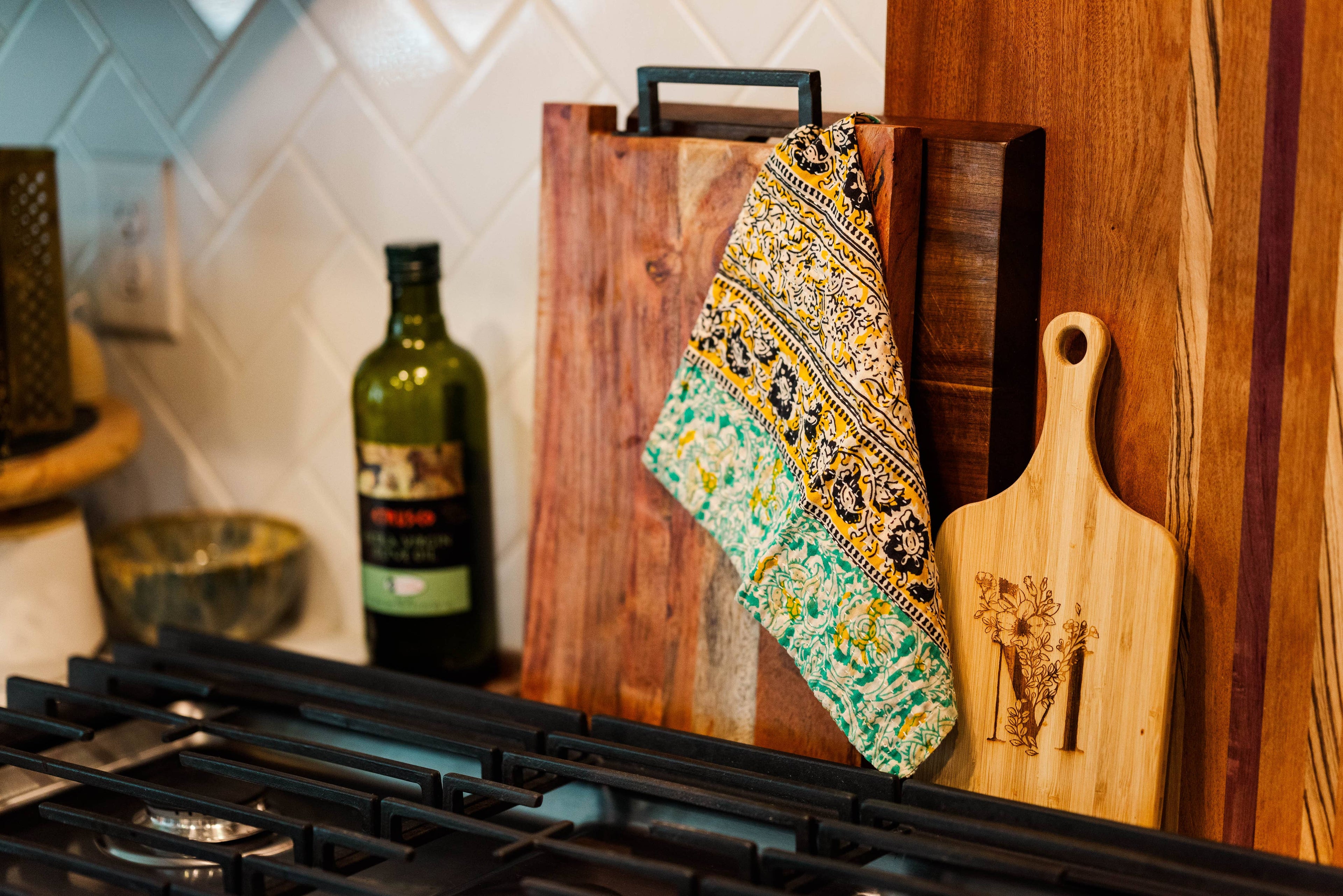 Wooden cutting board with a decorative towel and a bottle of olive oil on a kitchen counter.