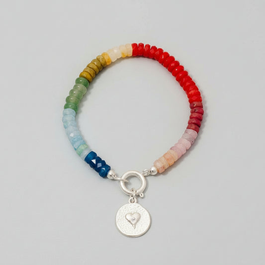 A bracelet with a silver heart coin charm and multicolored beads strung together, displayed on a beige textured surface.