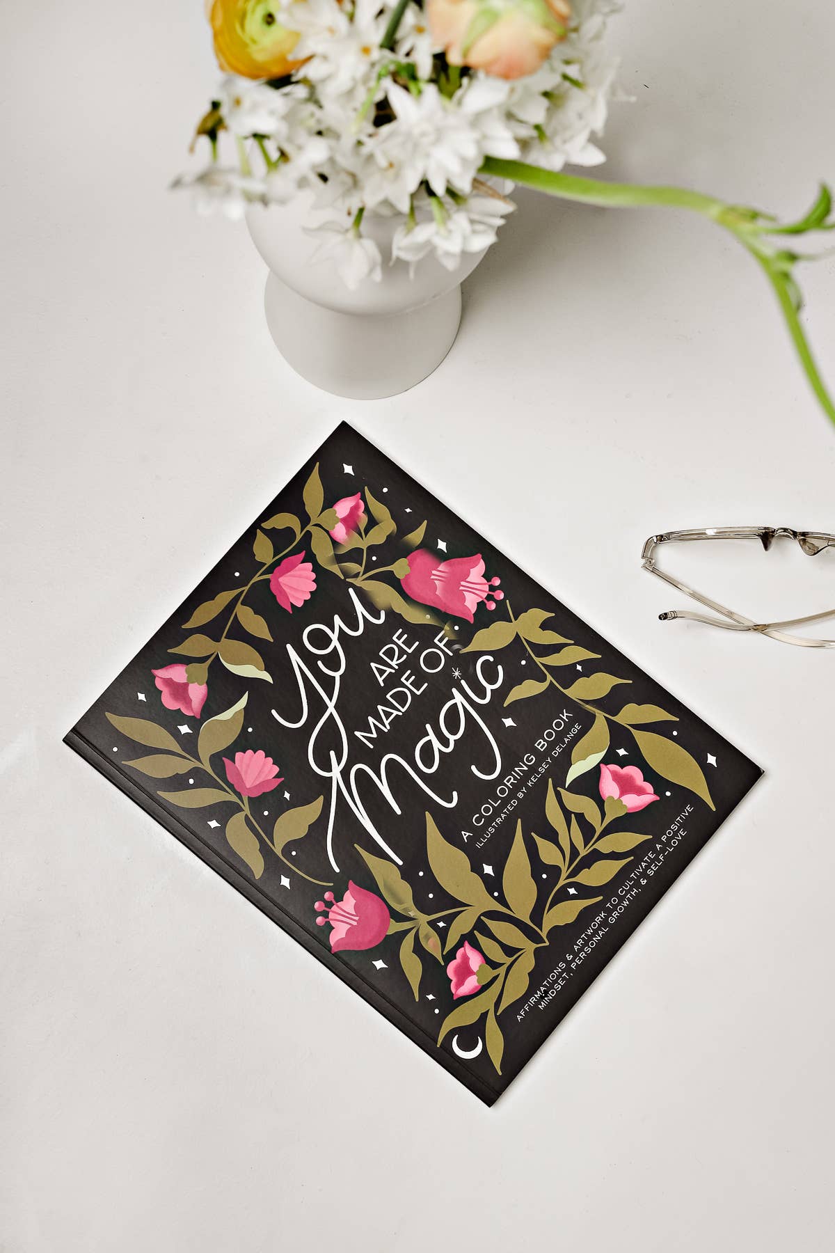Book titled 'You Are Made of Magic' with floral design on a white surface with flowers and glasses.