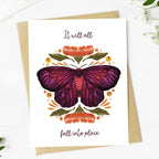 A greeting card with an illustration of a colorful moth surrounded by floral elements, and the text 'It will all fall into place' on the front. The card is accompanied by a kraft paper envelope and placed on a white surface with a subtle floral pattern in the background.