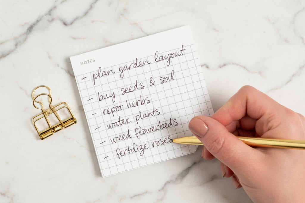 Handwritten to-do list on a small notepad with a gold pen and clip on a white background