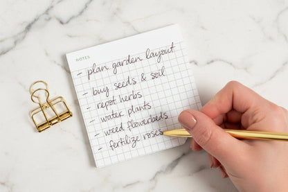 Handwritten to-do list on a small notepad with a gold pen and clip on a white background