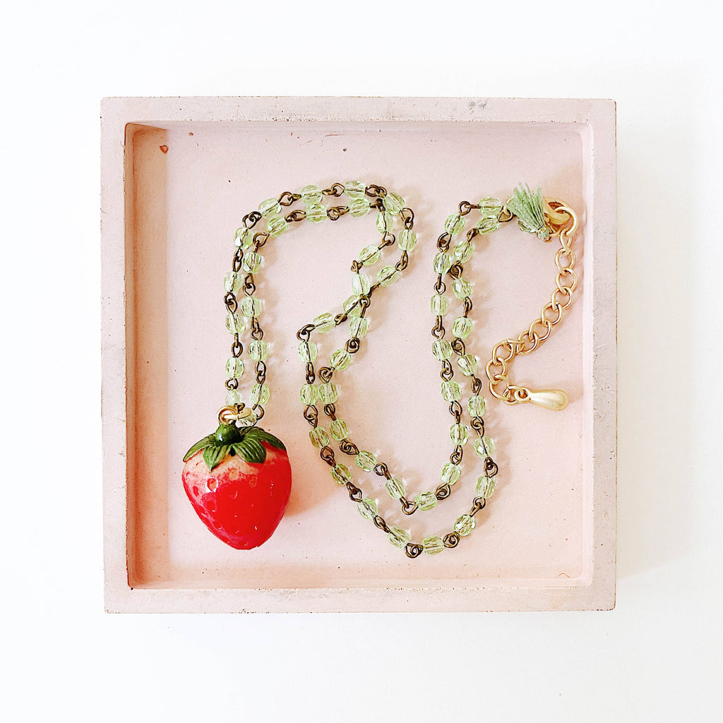 Overhead shot of strawberry necklace with lobster clasp and green tassel laid in a flat lay display