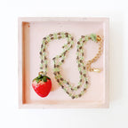 Overhead shot of strawberry necklace with lobster clasp and green tassel laid in a flat lay display