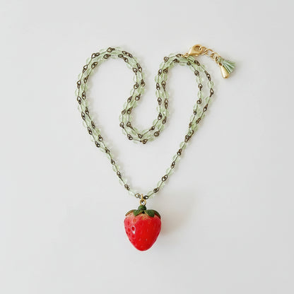 Overhead shot of strawberry necklace with lobster clasp and green tassel laid in a flat lay display