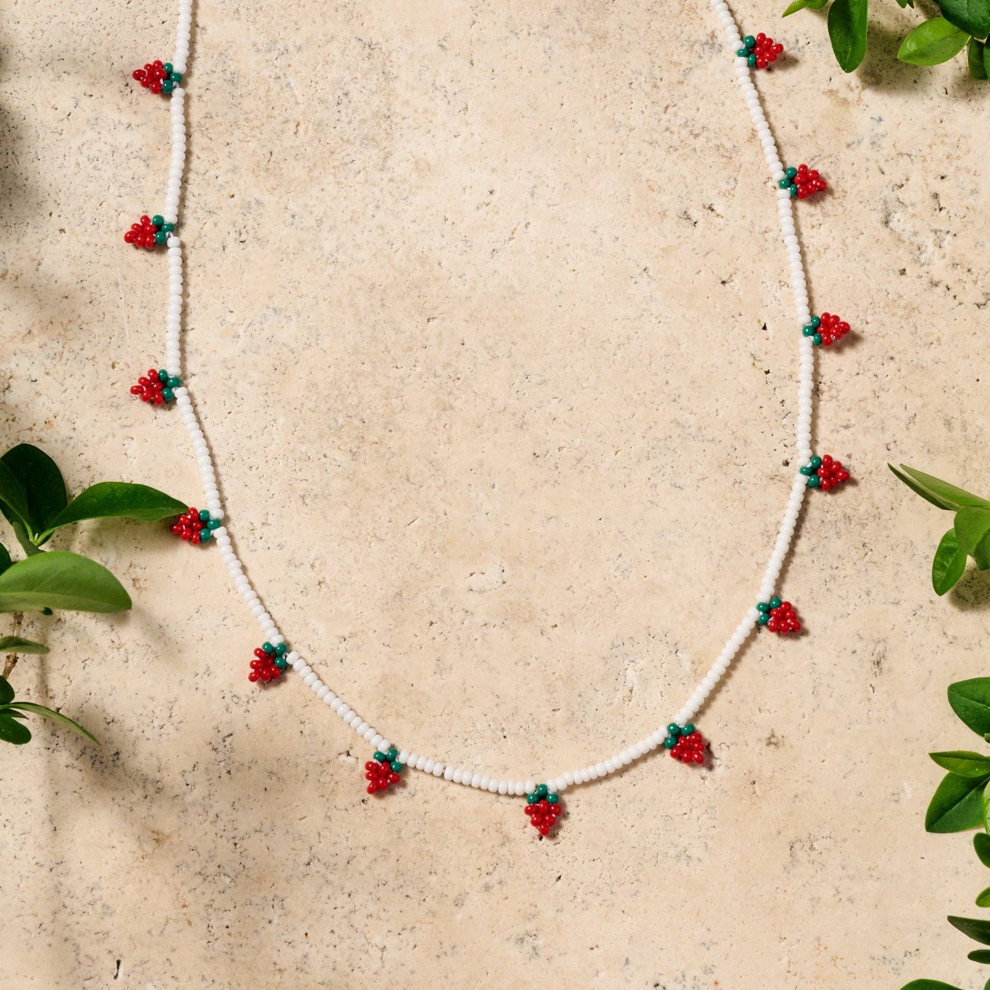 Delicate white glass bead necklace with red and green strawberry motif, hand-beaded on a stone surface