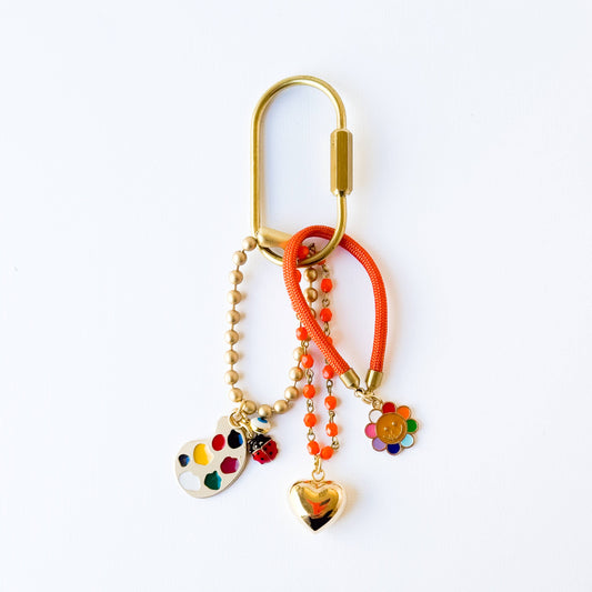 Close-up of keychain with gold heart, smiley face, rainbow, evil eye and ladybug
