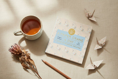 Teacup with tea, notebook titled 'White Crane a Day', pencil, and origami cranes on a textured surface