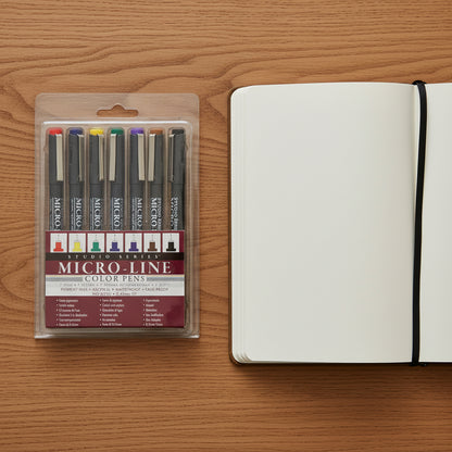 Studio Series Color Micro-Line Pen Set in clear case with 7 colored pens on a desk top next to an open journal