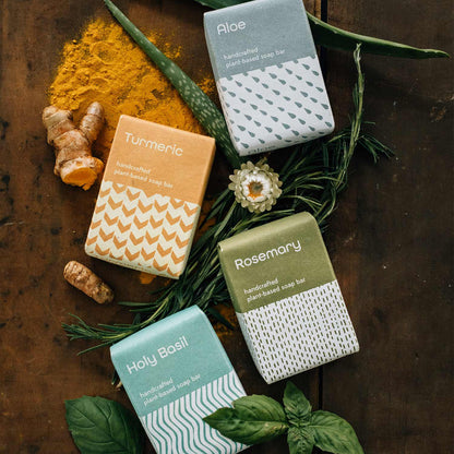 Four soap bars, including the aloe bar, with labels on a wooden surface with green leaves and herbs.