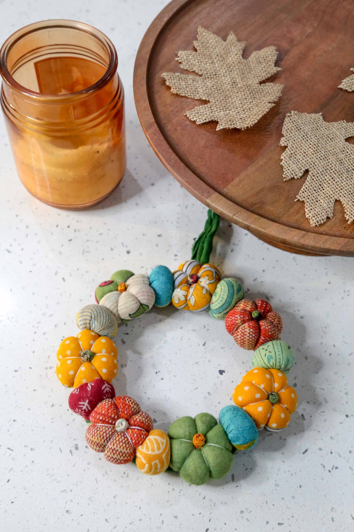Colorful felt pumpkin wreath on a white surface with a wooden tray and jar in the background.