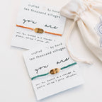 Two bracelets on cards with 'you are' message, 'crafted by hand ten thousand villages' text, and a drawstring bag.