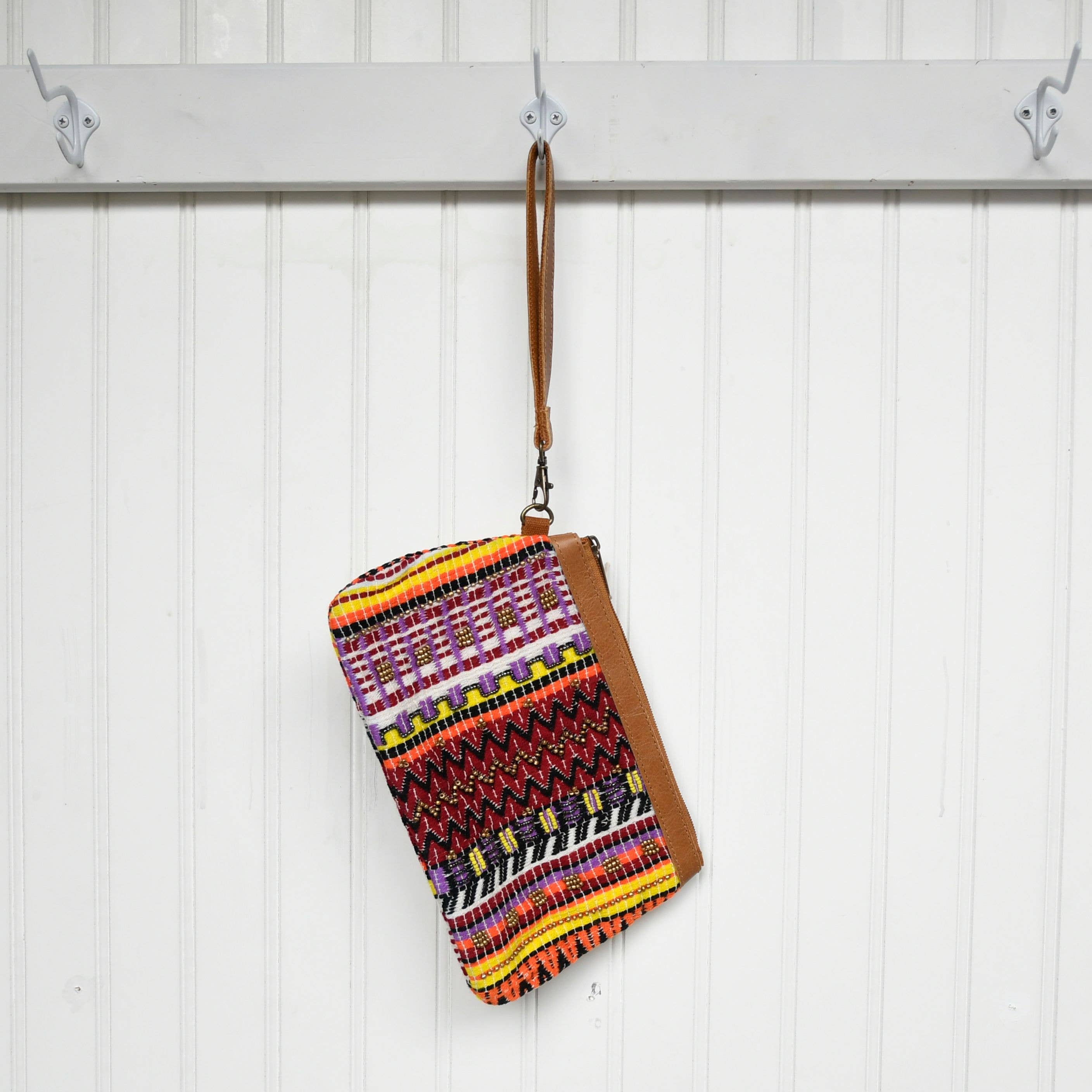 Colorful patterned pouch with a brown strap hanging on a white wooden panel.