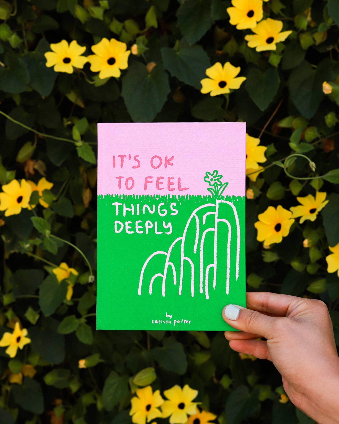 Hand holding a green and pink card with text and a plant illustration against a background of yellow flowers and green leaves.