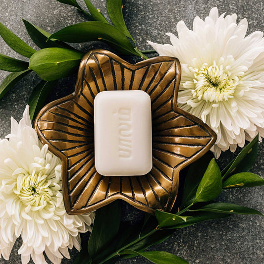 Bronze soap dish with a bar of soap on a dark surface with white flowers and green leaves.