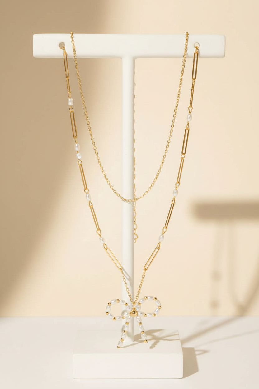 A gold layered chain necklace with a pearl beaded ribbon bow pendant on a white background.