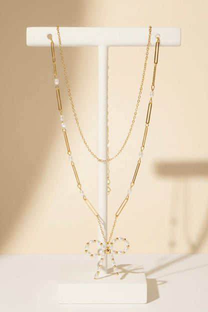 A gold layered chain necklace with a pearl beaded ribbon bow pendant on a white background.