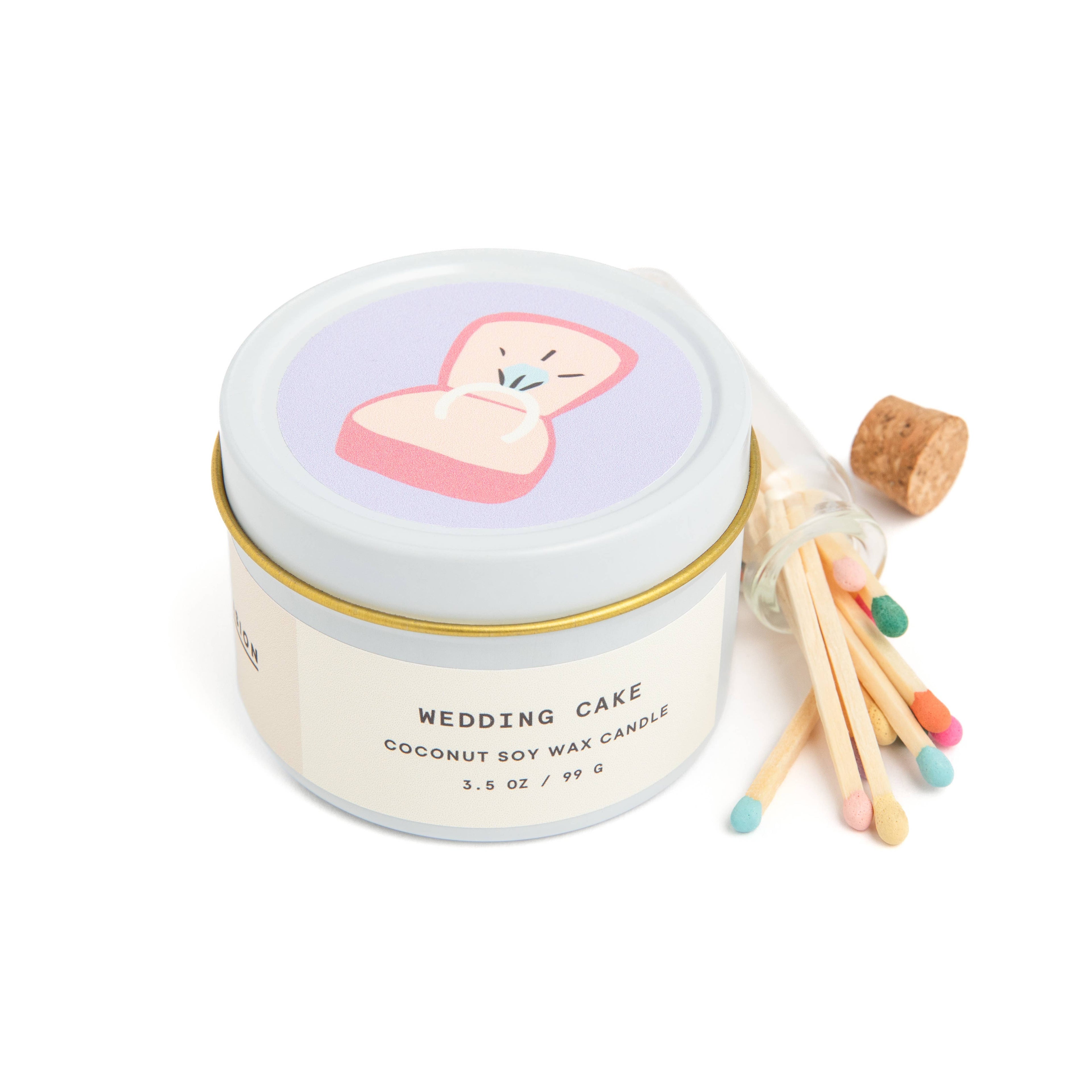 Wedding cake scented candle in white tin with corked vial of colorful matchsticks, packaged in a clear vinyl gift box.